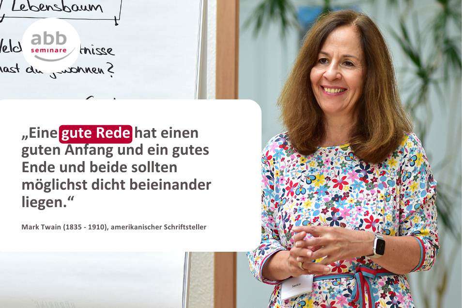 Das Bild zeigt eine lächelnde Frau mit schulterlangem braunen Haar, die vor einer Flipchart steht. Sie trägt ein bunt geblümtes Kleid und eine Smartwatch am Handgelenk. Ihre Hände sind in einer erklärenden Geste vor ihrem Körper. Im Hintergrund sind grüne Pflanzen zu sehen.

Links im Bild ist eine Flipchart mit handgeschriebenem Text erkennbar. Darüber ist das Logo von abb-seminare platziert.

Im unteren Bereich des Bildes befindet sich ein Zitat von Mark Twain in dunkler Schrift auf weißem Hintergrund:
„Eine gute Rede hat einen guten Anfang und ein gutes Ende und beide sollten möglichst dicht beieinander liegen.“
Die Worte „gute Rede“ sind rot hinterlegt und stechen hervor.

Das Bild visualisiert das Thema „richtig präsentieren“ und unterstreicht die Bedeutung einer klaren, prägnanten Rede.