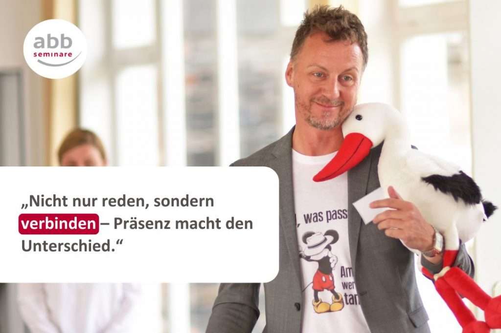 Das Bild zeigt einen Mann mit kurzen, leicht lockigen Haaren und einem freundlichen Lächeln. Er trägt ein graues Sakko über einem weißen T-Shirt mit einem Mickey-Mouse-Motiv. In seiner rechten Hand hält er einen Plüschstorch mit langem roten Schnabel und Beinen, der locker über seiner Schulter liegt.

Im Hintergrund ist eine unscharfe Person in weißer Kleidung zu erkennen. Der Raum ist hell und lichtdurchflutet, mit großen Fenstern im Hintergrund.

Links oben im Bild befindet sich das Logo von abb-seminare, bestehend aus einem weißen Kreis mit dem Schriftzug „abb seminare“ in Grau und Rot.

Zentral im unteren Bereich des Bildes steht ein Zitat in dunkler Schrift auf weißem Hintergrund:
„Nicht nur reden, sondern verbinden – Präsenz macht den Unterschied.“
Das Wort „verbinden“ ist hervorgehoben und mit einem roten Hintergrund hinterlegt.

Das Bild vermittelt die Bedeutung von Präsenz und Verbindung beim Präsentieren. Es zeigt, dass richtig präsentieren durch Kontakt mehr ist als nur Worte – es geht um echte Verbindung mit dem Publikum.